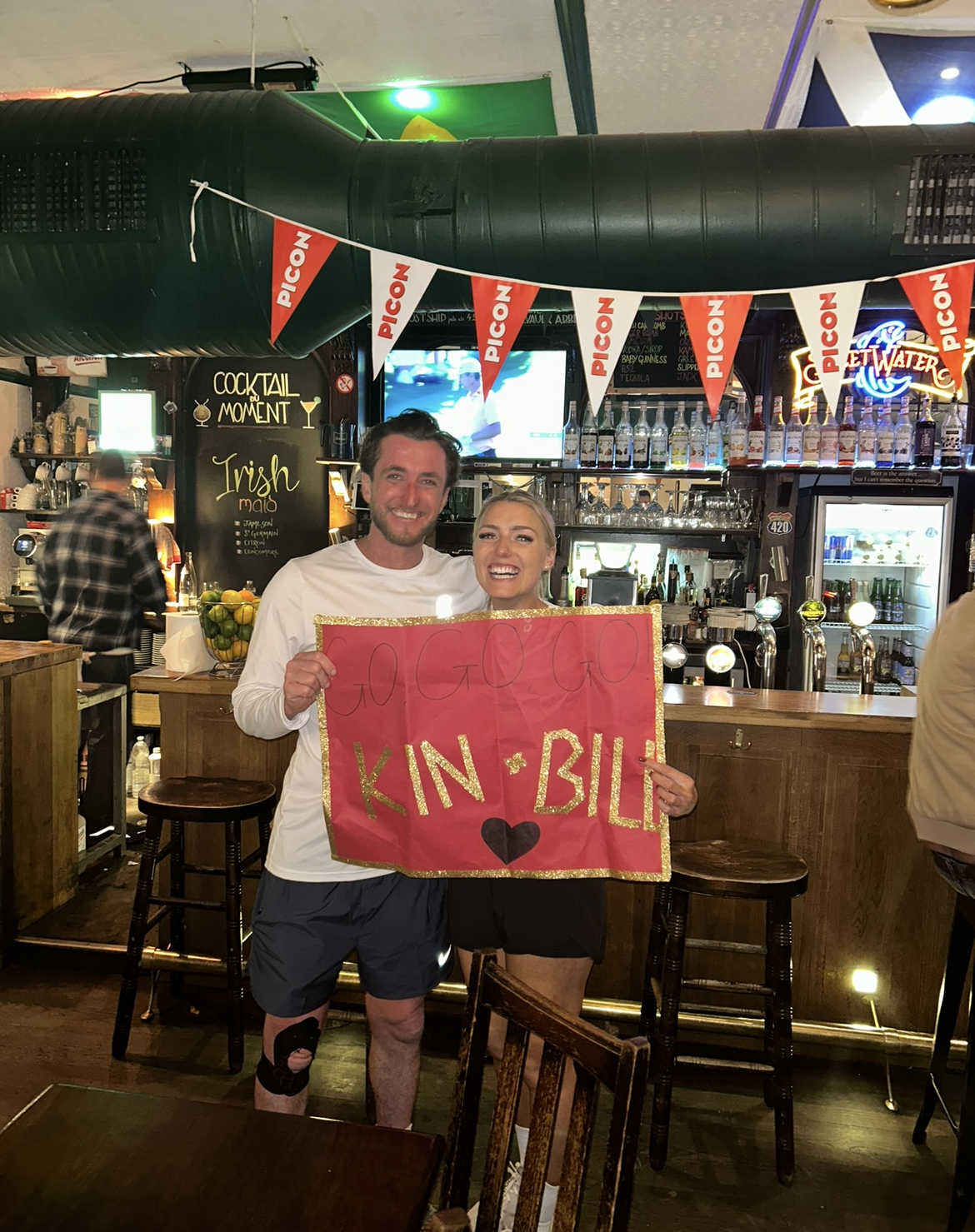 Two supporters holding a handmade 'Go Go Go Kin & Bill' sign in a Paris bar, cheering on the team.