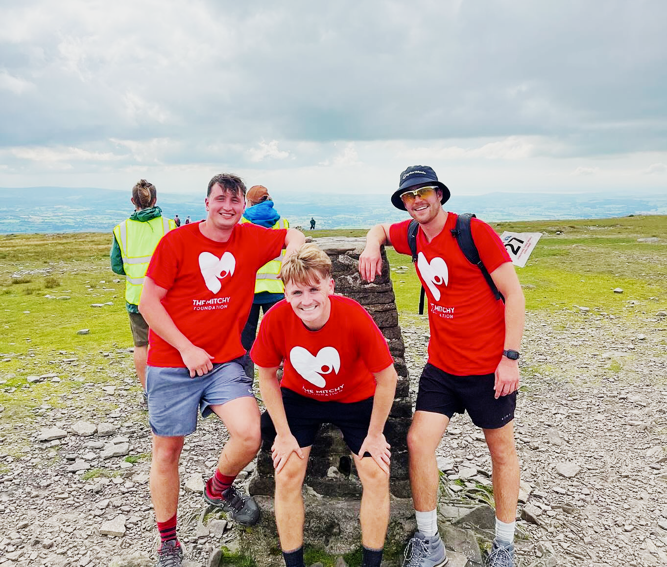 Three supporters in Mitchy Foundation shirts posing beside a trig point on the Yorkshire Three Peaks route.